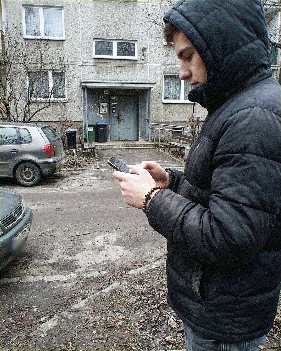 Syn w kurtce przed blokiem patrzy w telefon, na ręce bransoletka
